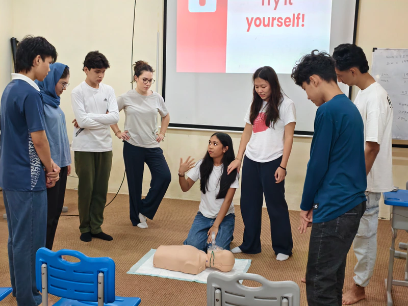 Workshop participants gather around a coach discussing CPR beside a training manikin