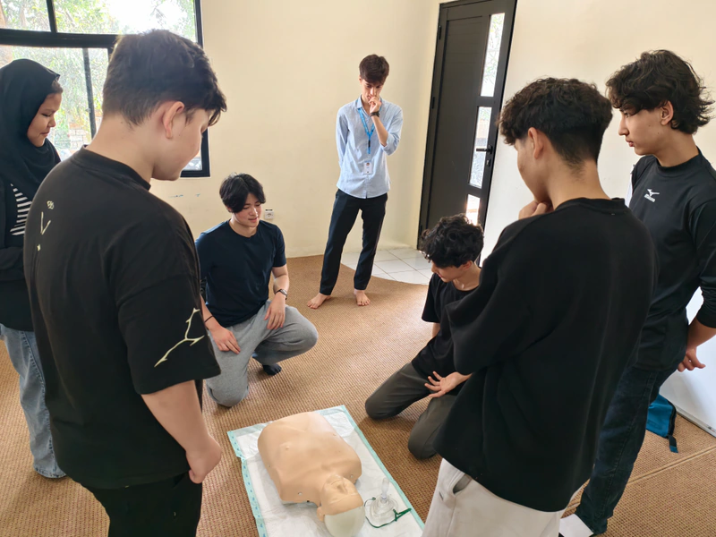 A small group of students gathers around a CPR manikin with a workshop coach