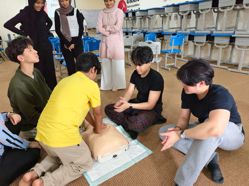 A participant in a yellow shirt practises chest compressions while coaches and classmates watch